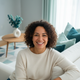A happy person in their late 20s or early 30s smiling warmly, sitting on a white sofa with teal and aqua pillows.